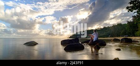 Langzeitbelichtung von Frauen, die bei Sonnenaufgang/Sonnenuntergang auf Felsen am Strand sitzen Stockfoto