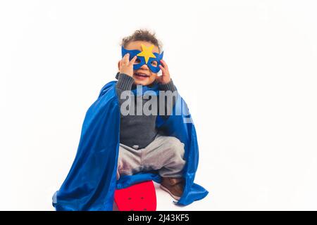 Niedlichen afroamerikanischen gewellten kleinen Jungen mit dem Kostüm eines Superhelden lächeln und berühren seine Maske Studio Schuss voller weißer Hintergrund Kopie Raum. Hochwertige Fotos Stockfoto