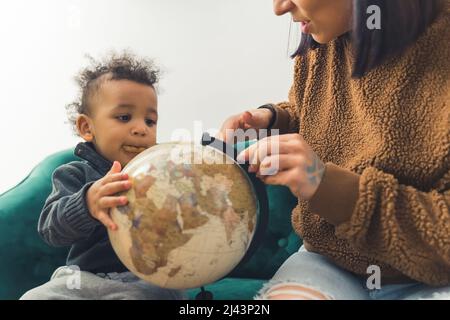 Süßer afroamerikanischer kleiner Junge mit einem Keks im Mund, der mit seiner Mutter auf dem grünen Sofa sitzt und den Globus anschaut, der von einem Cowboy gedreht wurde, das im Studio auf weißem Hintergrund einen Kopierraum geschossen hat. Hochwertige Fotos Stockfoto