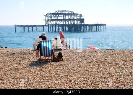 Urlauber in Liegestühlen genießen die Sonne vor dem verwelkenden Pier am Weststrand von Brighton in Brighton East Sussex England Stockfoto