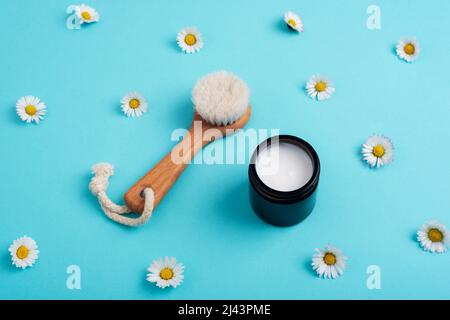 Holzpinsel und cremefarbenes Glas auf hellblauem Hintergrund mit Gänseblümchen. Hautpflege, natürliche hausgemachte Kosmetik-Konzept. Draufsicht. Stockfoto
