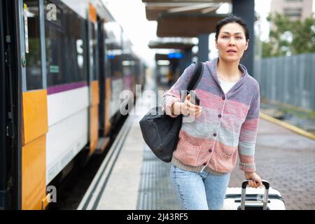 Eine Asiatin, die mit einem Rucksack und einem Koffer unterwegs ist, läuft am Bahnhofssteig entlang Stockfoto