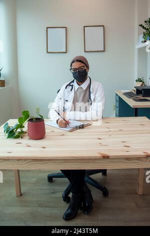 Ein Arzt in einer Maske sitzt lächelnd in ihrem Büro und macht Papierkram Stockfoto