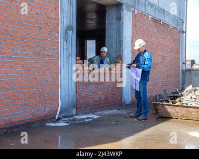 Kemerowo, Russland - 24. juni 2021. Der Vorarbeiter mit den Konstruktionszeichnungen prüft die Qualität der Arbeit des Maurers, der die Scheidewände des Rahmengebäudes aufbaut Stockfoto