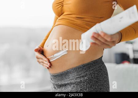 Hautpflege während der Schwangerschaft. Schwangerin, die Creme auf den Bauch für Dehnungsstreifen auftragend. Feuchtigkeitsspendende Nahaufnahme der Hände und Lotion Flasche zu Hause Stockfoto