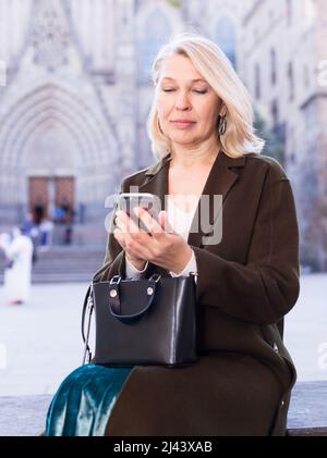 Elegante Frau, die auf den Bildschirm eines Mobiltelefons schaut, das auf einer Straßenbank sitzt Stockfoto