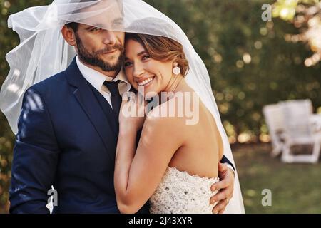 Ich bin so froh, dass ich ihn geheiratet habe. Ausgeschnittene Aufnahme einer anhänglichen jungen Braut, die am Hochzeitstag lächelt und ihren Bräutigam unter einem Schleier umarmt. Stockfoto