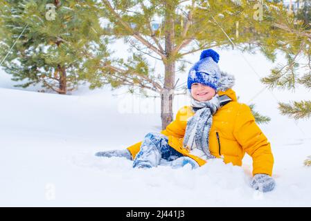 Fröhlicher Junge spielt Schneebälle in einer gelben Jacke und einem blauen Hut Stockfoto