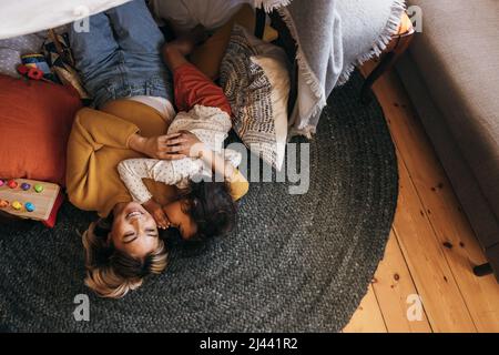 Draufsicht auf ein junges Mädchen, das spielerisch in das Ohr ihrer Mutter flüstert. Fröhliche Mutter und Tochter liegen auf dem Boden in ihrem Spielbereich. Mutter und da Stockfoto