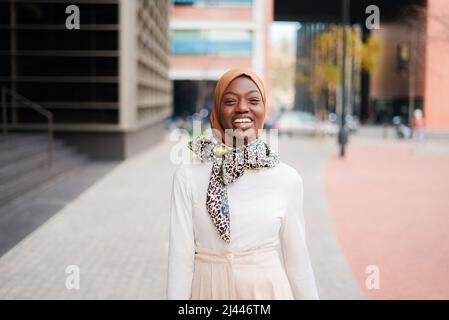 Positive, gut gekleidete afroamerikanische Frau im islamischen Kopftuch, die im modernen Stadtteil steht und mit einem breiten Lächeln auf die Kamera blickt Stockfoto