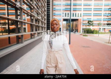 Selbstbewusste afroamerikanische Frau in modernem Outfit und muslimischer Hijab, die sich gegen moderne Wolkenkratzer im Business Center auflehnen und die Kamera betrachten Stockfoto