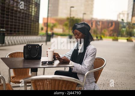 Fokussierte afroamerikanische Frau in eleganter Freizeitkleidung und traditionellem islamischen Kopftuch, die bei einer Tasse Kaffee am Tisch sitzt und im Mobiltelefon surft Stockfoto