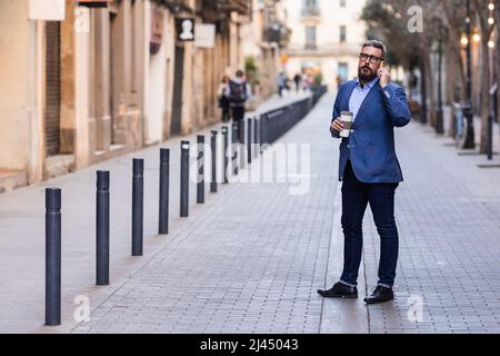 Gut gekleideter Mann, der in der Innenstadt steht und telefoniert und die Kaffeetasse hält Stockfoto