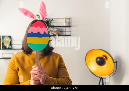 Junge dunkelhaarige Mann trägt gefälschte Kaninchenohren versteckt Gesicht hinter dem Papier Osterei Figur auf einem Stick Ostern Konzept Medium Nahaufnahme kopieren Raum Hause Hintergrund . Hochwertige Fotos Stockfoto