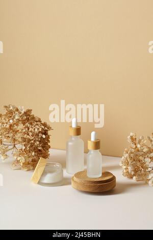 Transparente Tropf-Kosmetikflaschen und ein Glas Feuchtigkeitscreme mit trockenen Blüten auf beigem Hintergrund. Design von Naturkosmetischen Produkten Stockfoto