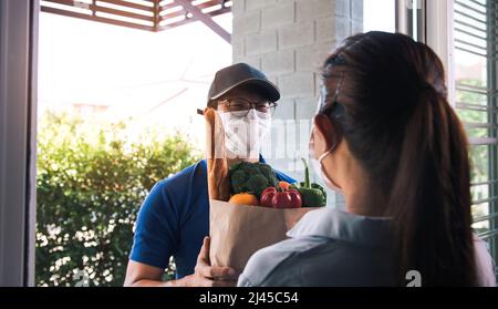 Lieferung eines asiatischen Mannes mit Schutzmaske während einer Epidemie Virus, während er eine Tüte mit Gemüse Lebensmittel an eine weibliche Kundin an der d Stockfoto