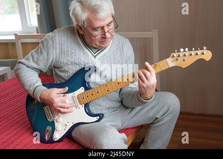 Älterer Mann spielt Gitarre. Älterer Mann, der auf dem Sofa sitzt und Gitarre spielt. Porträt eines grauhaarigen reifen Mannes mit Brille, der das Spielen lernt. Stockfoto