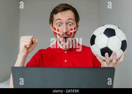 Junge aufgeregt Mann Fan unterstützen Dänemark Fußballnationalmannschaft halten in der Hand Fußball Fernsehen Live-Stream auf Laptop. Stockfoto