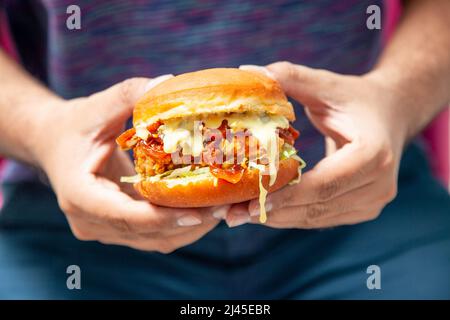 Mann mit einem würzigen gebratenen Hähnchenburger, der mit geschmolzenem Käse übergossen wird. Stockfoto