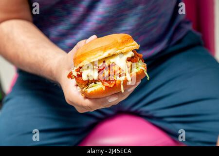 Mann mit einem würzigen gebratenen Hähnchenburger, der mit geschmolzenem Käse übergossen wird. Stockfoto