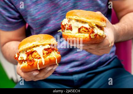 Mann mit zwei scharf gebratenen Hühnerburgern, die mit geschmolzenem Käse übergossen werden. Stockfoto