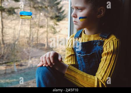 Weltweite Krise der ukrainischen Geopolitik. Unglückliches kleines Mädchen, das allein auf dem Fensterbrett zu Hause sitzt. Traurigkeit sehnsüchtige Hoffnung. Zeichen der ukrainischen Flagge auf Kind Stockfoto