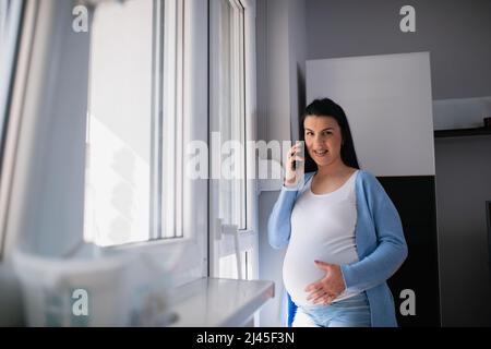 Eine schwanger kaukasische Frau benutzt ein Mobiltelefon, spricht, lächelt und blickt direkt auf die Kamera, während sie neben der Tür des Wohnzimmers steht. Stockfoto