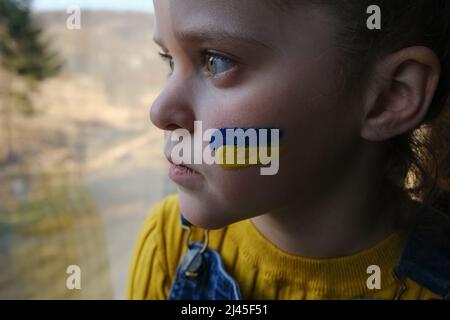 Zeichen der ukrainischen Flagge auf der Wange des Kindes. Weltweite Krise der ukrainischen Geopolitik. Unglückliches kleines Mädchen, das allein auf dem Fensterbrett zu Hause sitzt. Lange Traurigkeit Stockfoto