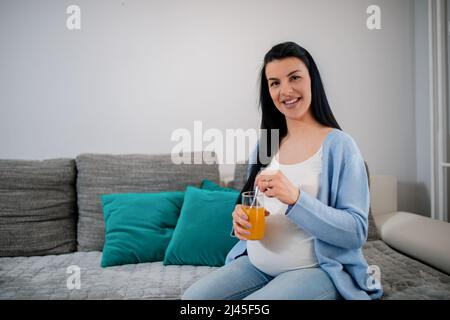 Die hübsche kaukasische Schwangerin sitzt auf dem Sofa und mischt frischen Fruchtsaft mit einem Löffel. Schwarz haarige Frau mit schwanger Bauch sieht direkt an der Kamera. Stockfoto