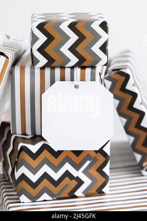 Nahaufnahme mit leerem Geschenkanhänger. Geschenkschachteln in geometrischem Papier mit Streifen- und Chevron-Muster verpackt. Weihnachten, Neujahr, Geburtstag, Jubiläum Label Mock Stockfoto