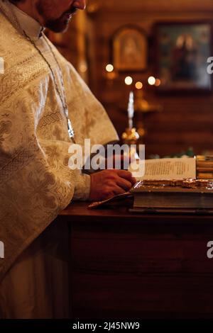 Beschnittenes Foto des Priesters tragen zeremonielle Soutane, die an einem Rednerpult mit Büchern steht und das Gebetsbuch in der orthodoxen Kirche liest. Stockfoto