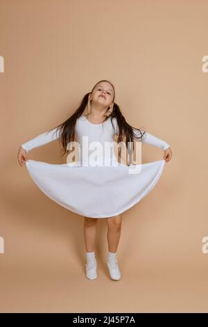 Junge niedliche lächelnde Mädchen mit langen dunklen Haaren in weißem Kleid, Socken und Turnschuhe stehen, halten Kleid mit Händen, Spitze-Toeing, Spaß an Stockfoto