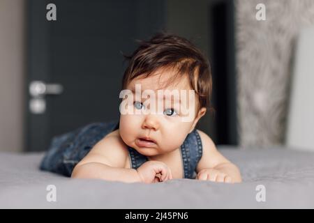 Portrait eines hübschen Babys, das Denim-Strampler trägt und zu Hause auf einer grauen Decke liegt. Verwirrt aussehendes Kleinkind kriecht im Schlafzimmer auf dem Bett, erkunden Sie die Welt Stockfoto