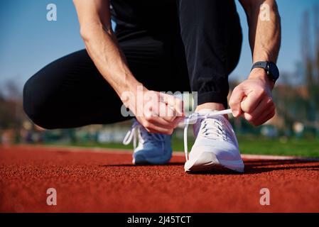 Männlicher Läufer in weißen Sneakers mach dich bereit für den Lauf auf der Stadionstrecke. Männliche Hände binden an Sport-Sneakers zum Joggen. Fitness und gesunder Lebensstil Stockfoto
