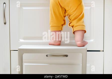 Kleinkind Baby Junge kletterte mit seinen Füßen auf einem Hochstuhl auf den Küchenschrank klettern. Probleme mit der Kindersicherheit im heimischen Zimmer, kleines Kind Stockfoto