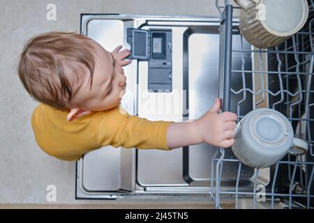 Baby Junge Kleinkind steht bei einer offenen Spülmaschine mit schmutzigem Geschirr im Inneren. Probleme mit der Kindersicherheit im heimischen Zimmer, kleines Kind Stockfoto