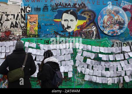 Der russische Präsident Wladimir Putin wird auf der Lennon-Mauer (Lennonova zeď) in Prag, Tschechien, dargestellt. Präsident Wladimir Putin wird mit charakteristischem Haarschnitt und Zahnbürstenbart von Adolf Hitler dargestellt, der auf der blutigen Karte der Ukraine platziert wurde, um gegen die russische Invasion in der Ukraine im Jahr 2022 zu protestieren. Die tschechische Inschrift über Putins Kopf „Válka je vůl“ (literarisches Mittel: Krieg ist ein Ochse) ist eine Zeile aus einem beliebten Lied der tschechischen Rockband „Synkopy 61“. Touristen, die am 2. April 2022 abgebildet wurden, lasen Antikriegstexte, die neben der Lennon-Mauer platziert wurden. Stockfoto