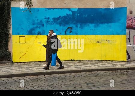 Fußgänger kommen an der ukrainischen Nationalflagge vorbei, die auf der Mauer in der Karmelitská-Straße auf der Kleinseite in Prag, Tschechien, abgebildet am 2. April 2022, abgebildet ist. Die riesige Flagge wurde im historischen Zentrum von Prag dargestellt, um ukrainische Flüchtlinge in der Tschechischen Republik zu unterstützen und gegen die russische Invasion in der Ukraine im Jahr 2022 zu protestieren. Stockfoto