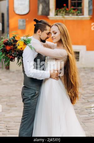 Schönes junges Paar, das sich im Hof auf einem Hintergrund eines orangefarbenen Gebäudes umarmt. Charmantes Mädchen mit langen Haaren Stockfoto