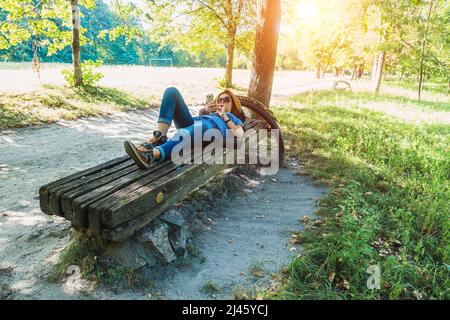 Ein Mädchen liegt mit einem Smartphone auf einer Straßenbank in einem Park. Stockfoto