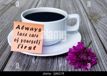 Text auf Stocknotiz mit weißer Kaffeetasse und violetter Blume auf Holzschreibtisch - kümmern Sie sich immer zuerst um sich selbst. Stockfoto