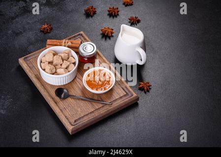 Köstliche Aprikosenmarmelade, Milch und Zucker auf einem Holzschneidebrett vor dunklem Betonboden Stockfoto