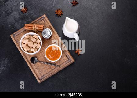 Köstliche Aprikosenmarmelade, Milch und Zucker auf einem Holzschneidebrett vor dunklem Betonboden Stockfoto