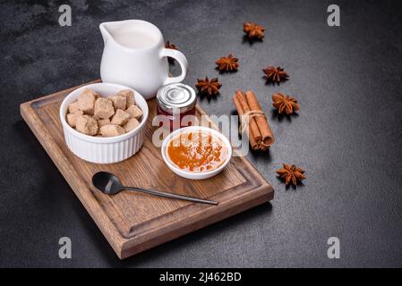 Köstliche Aprikosenmarmelade, Milch und Zucker auf einem Holzschneidebrett vor dunklem Betonboden Stockfoto