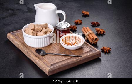 Köstliche Aprikosenmarmelade, Milch und Zucker auf einem Holzschneidebrett vor dunklem Betonboden Stockfoto