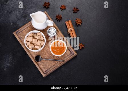 Köstliche Aprikosenmarmelade, Milch und Zucker auf einem Holzschneidebrett vor dunklem Betonboden Stockfoto