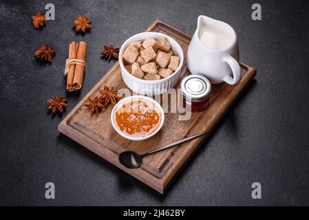 Köstliche Aprikosenmarmelade, Milch und Zucker auf einem Holzschneidebrett vor dunklem Betonboden Stockfoto