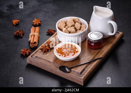 Köstliche Aprikosenmarmelade, Milch und Zucker auf einem Holzschneidebrett vor dunklem Betonboden Stockfoto