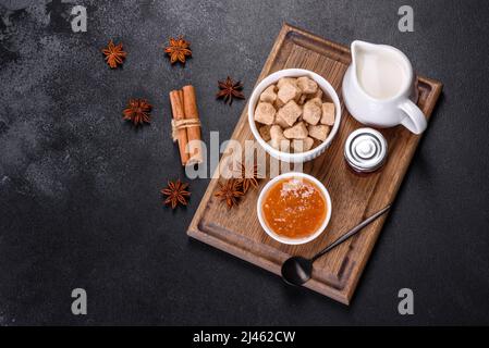Köstliche Aprikosenmarmelade, Milch und Zucker auf einem Holzschneidebrett vor dunklem Betonboden Stockfoto