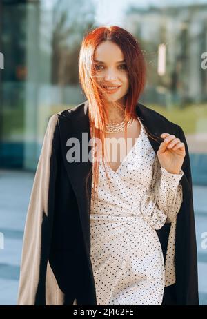 Porträt der stilvollen rothaarigen Frau in weißem Kleid und schwarzer Jacke auf Bürogebäude Hintergrund Stockfoto